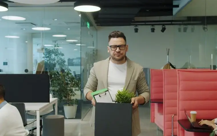 A man in glasses walking out of an office holding a box of personal belongings and a potted plant