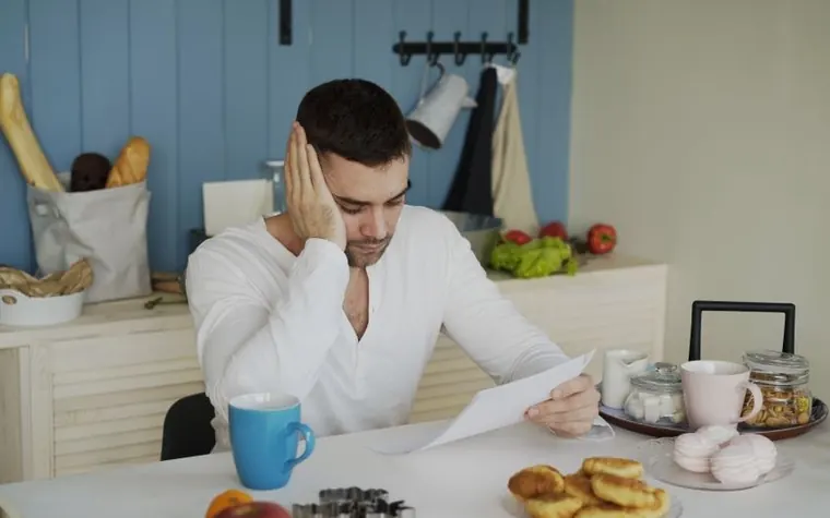 A man resting his cheek on one hand while reading a paper document at a kitchen table