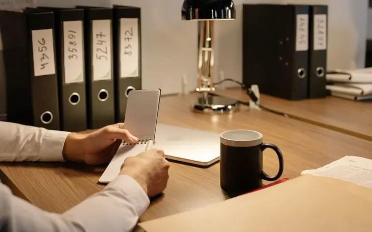 A person at an office desk holding a notebook beside a coffee mug