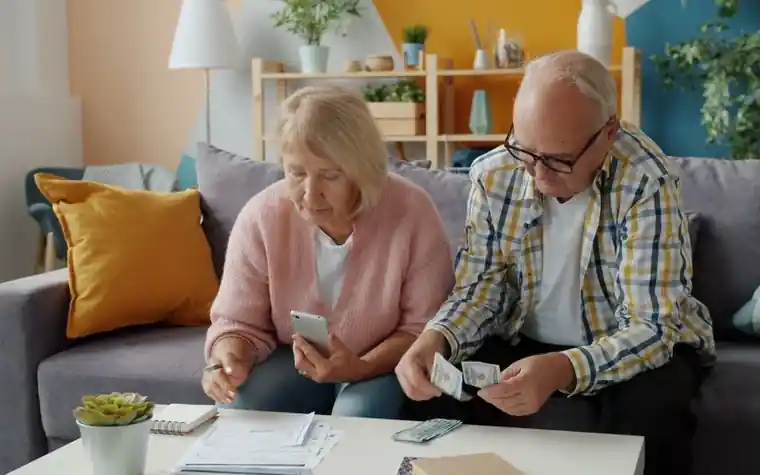 An elderly couple sitting on a sofa, talking while looking at documents and a phone