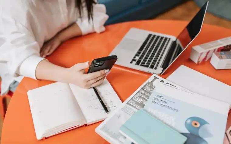 A smartphone in hand over a table with a notebook and laptop spread out