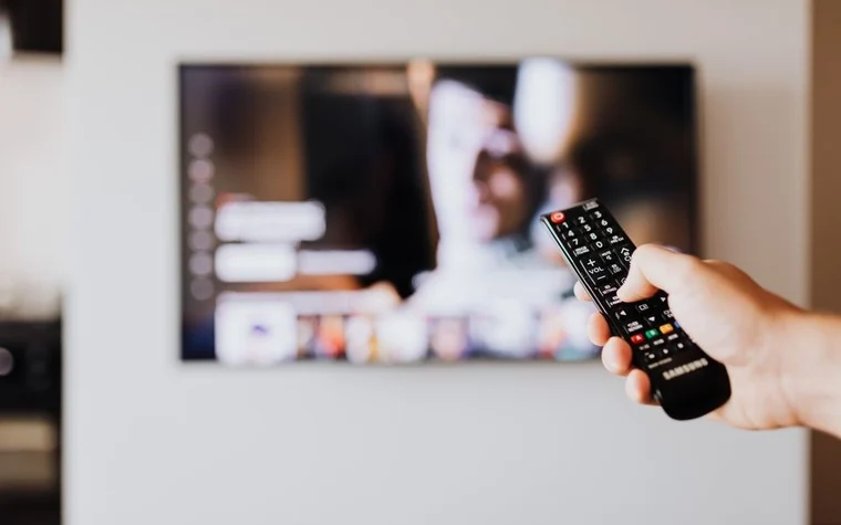 A hand holding a TV remote with a blurred television screen in the background