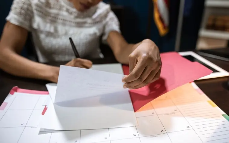 A hand writing a schedule with a pen on a calendar and papers spread across a desk