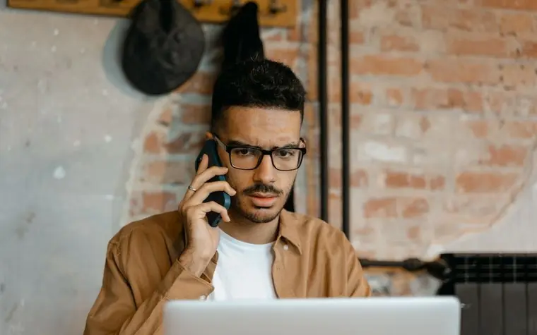 A man with glasses talking on a mobile phone in front of a laptop