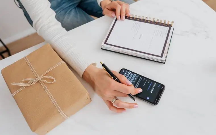 A person at a white table looking at a smartphone with one hand and taking notes in a notebook with the other